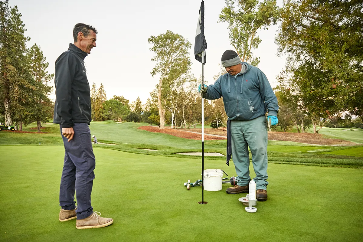 A golf course superintendent looks on as a crew member adjusts a cup cutter on a putting green at sunrise.