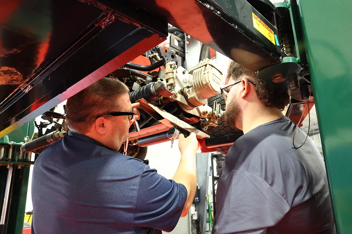 Two equipment technicians in safety glasses service the underside of a fairway mower in a maintenance shop.